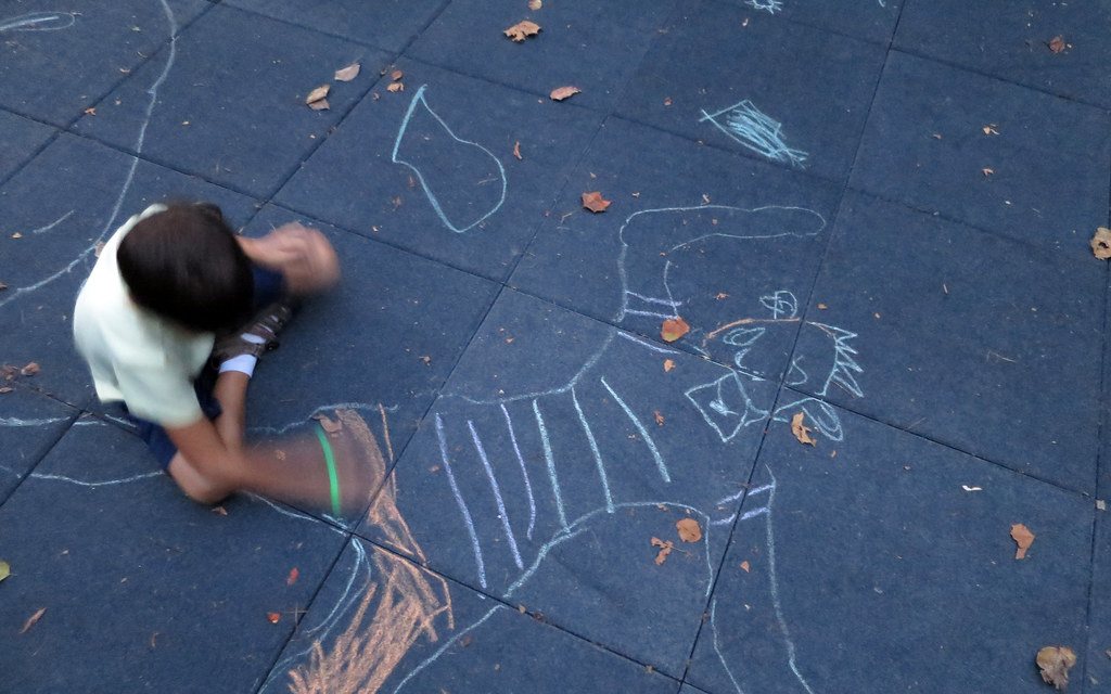A young child drawing a large chalk picture on pavement